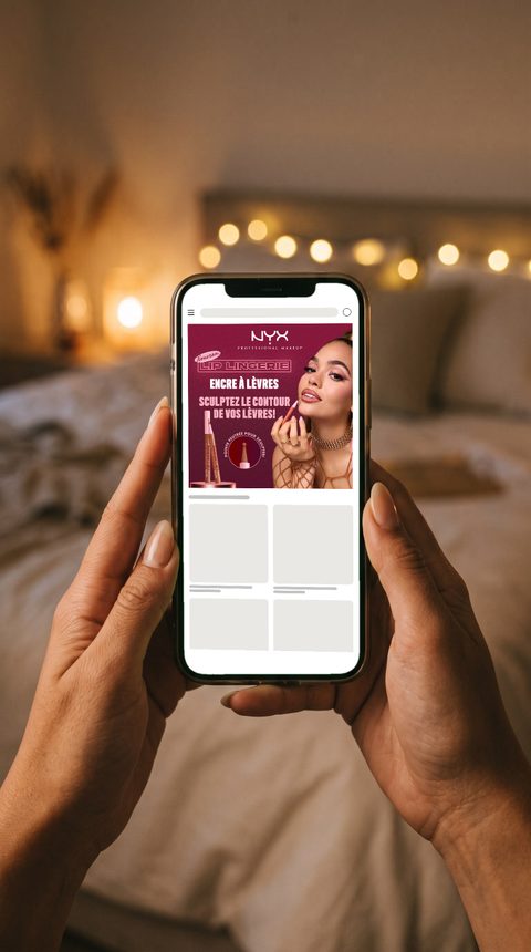 Mise en situation : bannière NYX Lip Lingerie « Encre à lèvres » sur un téléphone tenu à deux mains, dans une chambre éclairée par une guirlande.