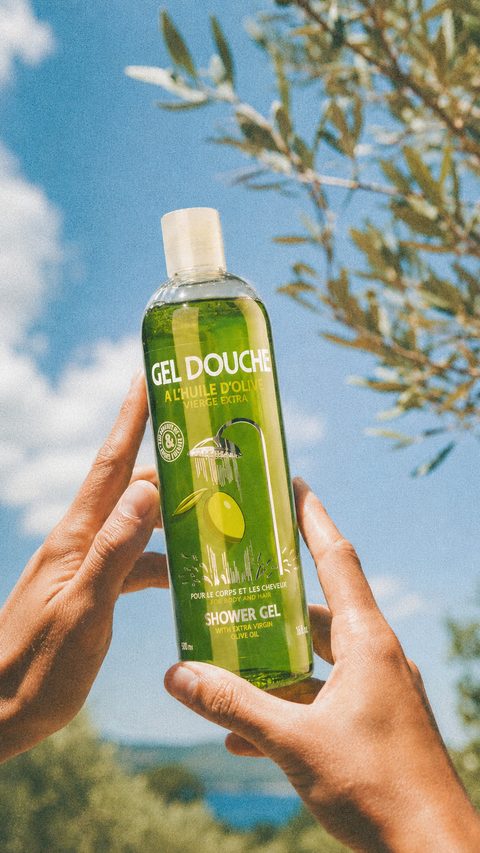 Une main tient une bouteille de gel douche Une Olive en Provence devant un olivier et la mer.